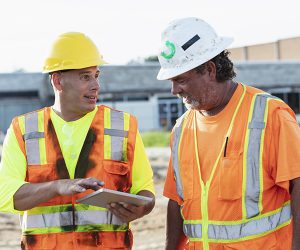 Two mature men working at a construction site, conversing and looking at a digital tablet.